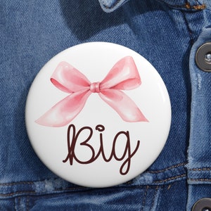 May include: A white button with a pink watercolor bow and the word "big" written in brown cursive.