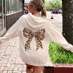 Puede incluir: Una sudadera con capucha de color crema con un gran lazo de estampado de leopardo en la espalda.