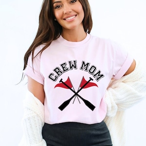 May include: A pink t-shirt with the text "CREW MOM" in black letters with a white outline. The text is surrounded by two crossed oars with red flags.