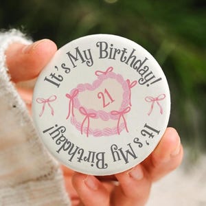May include: A circular white button featuring the text It's My Birthday! in dark grey lettering. The center displays a pink heart-shaped cake illustration with the number 21 and decorative pink bows surrounding the cake design.