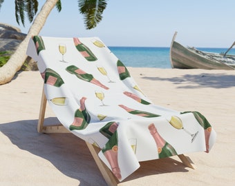 May include: A white beach towel with a repeating pattern of champagne bottles and glasses. The towel is draped over a wooden beach chair on a sandy beach.