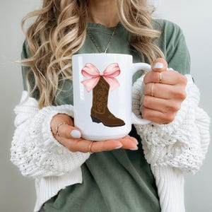 May include: White ceramic mug with a brown cowboy boot with a pink bow on top.
