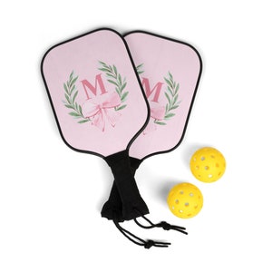 Könnte beinhalten: Zwei rosa Pickleball-Schläger mit einem floralen Kranz und dem Buchstaben "M" in der Mitte. Die Schläger haben schwarze Griffe und sind mit einer schwarzen Schnur zusammengebunden. Zwei gelbe Pickleball-Bälle sind enthalten.