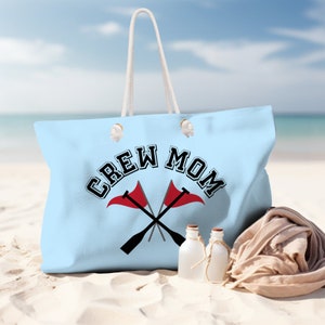 Puede incluir: Bolsa de lona azul claro con asas de cuerda blanca. La bolsa tiene el texto "CREW MOM" en negro con un gráfico de dos remos cruzados y banderas rojas.