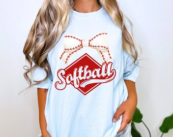 May include: A light blue short-sleeve t-shirt featuring a graphic print of a white bow with red baseball-style stitching. Below the bow is a red diamond shape containing the word Softball in a red cursive script font.