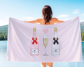 May include: A light pink beach towel featuring illustrations of two champagne bottles, a champagne glass, and a pink heart. The bottles have gold tops and red and black bows. The towel is held up by a person in front of a lake.