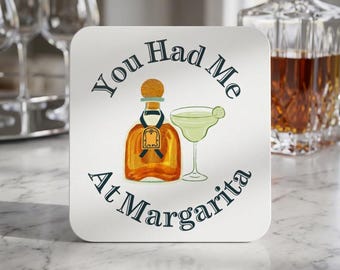 Può includere: Un sottobicchiere quadrato bianco con un'illustrazione centrale di una bottiglia di tequila color ambra e un bicchiere da margarita verde chiaro con una fetta di lime. Il testo "You Had Me At Margarita" è stampato in blu scuro attorno.