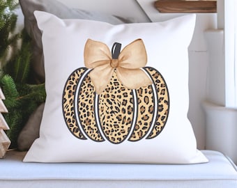 Puede incluir: Cojín cuadrado blanco con un diseño de calabaza. La calabaza tiene un estampado de leopardo en tonos marrón y beige, con un lazo beige en la parte superior. El cojín es un acento decorativo para el otoño.