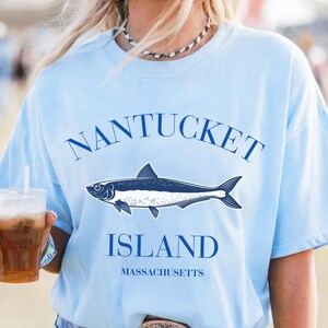 Könnte beinhalten: Hellblaues T-Shirt mit dem Schriftzug "NANTUCKET ISLAND MASSACHUSETTS" in Marineblau. Eine marineblaue Fischillustration befindet sich unter dem Wort "NANTUCKET". Das Shirt ist ein lässiger Stil.