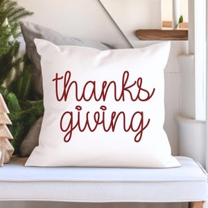 Peut inclure: Coussin carré blanc avec l'inscription "thanks giving" en script bordeaux. Le coussin est posé sur une surface blanche, avec un sapin de Noël et une décoration d'arbre en bois en arrière-plan.