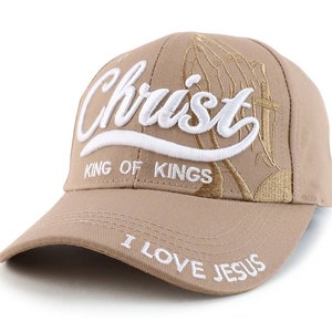 May include: A tan baseball cap with white embroidery. The front of the cap says "Christ King of Kings" with a praying hands design. The back of the cap says "I Love Jesus".