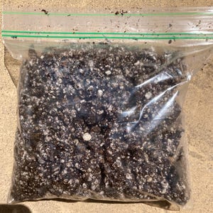 May include: A clear plastic bag filled with dark brown soil and white particles. The bag is sealed with a green zipper closure. The soil appears to be a potting mix, likely for plants.