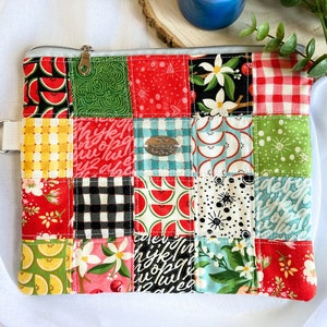 May include: A patchwork zippered pouch with a variety of colorful fabrics, including red, green, yellow, blue, and black. The pouch has a zipper closure and a small fabric loop on the side. The fabric patterns include watermelon slices, flowers, and geometric designs.