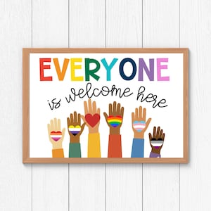 Puede incluir: Un cartel blanco con marco de madera dice "EVERYONE is welcome here" en letras de colores. Siete manos coloridas con corazones están levantadas en el aire, cada corazón mostrando una bandera del orgullo diferente.