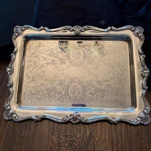 May include: A rectangular silver serving tray with ornate, scrolling edges and a detailed floral pattern. The tray is highly reflective, with a polished finish. The tray is likely made of silver-plated metal and is suitable for serving food or displaying decorative items.