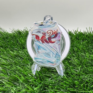 May include: A crushed Diet Coke can ornament with the Diet Coke logo in red and white. The ornament is displayed on a clear stand against a backdrop of green artificial grass.