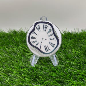 May include: A clear, melted-style clock with a white face, black Roman numerals, and black hands. The clock is mounted on a clear plastic stand, set against a backdrop of green artificial grass.
