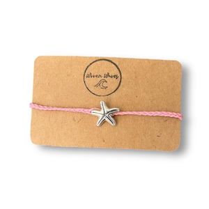 May include: A pink string bracelet with a silver starfish charm, presented on a brown card. The card features a circular logo with the text "Woven Waves" and a wave graphic. This bracelet is a simple, beach-themed accessory.