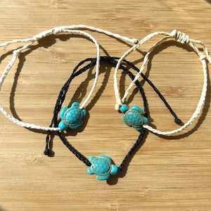 May include: Three adjustable bracelets with turquoise turtle charms. One bracelet is white, one is black, and one is off-white. The bracelets are on a wooden surface.