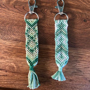 May include: Two handmade keychains with woven green and white patterns. Each keychain has a silver clasp and a tassel at the bottom. The keychains are displayed on a wooden surface.