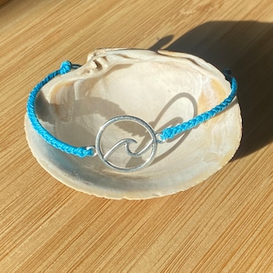 May include: A blue braided bracelet with a silver charm featuring a wave design. The bracelet is displayed on a seashell.