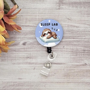 May include: A blue and white badge reel featuring a cartoon sloth sleeping on a pillow. The badge reel has the text "SLEEP LAB zzz" on it.