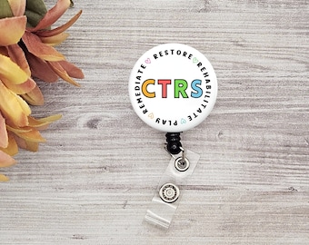 Op de afbeelding: Wit rond badge met een zwarte intrekbare badge-rol. Het badge heeft de tekst "CTRS" in kleurrijke letters omringd door de woorden "Remediate, Play, Rehabilitate, Restore" in een cirkel.