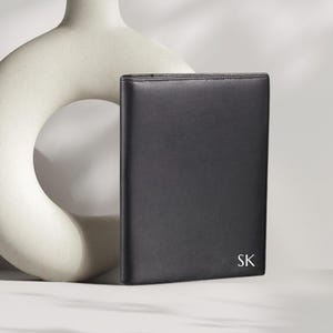 May include: A black leather portfolio with the initials "SK" in white. The portfolio is rectangular and appears to be closed. A cream-colored, abstract ceramic vase is in the background.