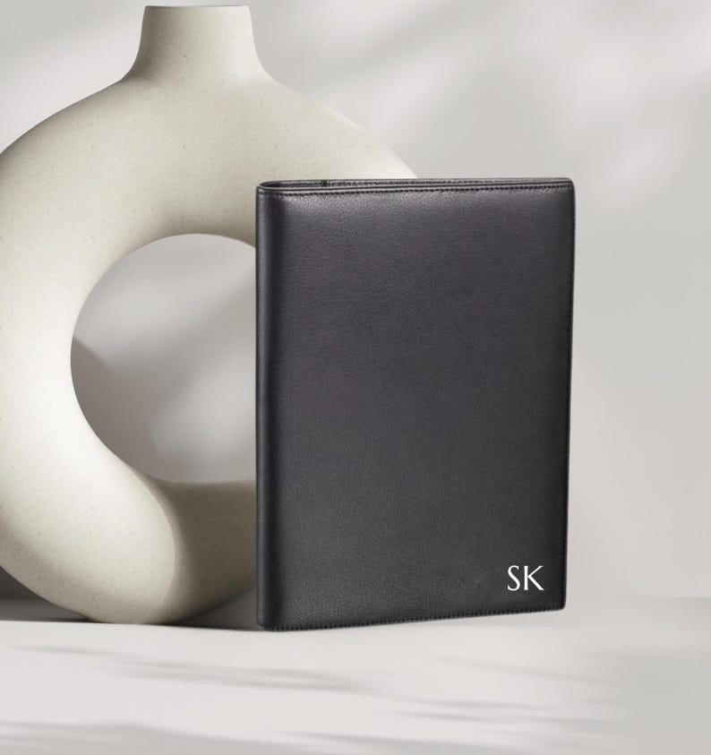 May include: A black leather portfolio with the initials "SK" in white. The portfolio is rectangular and appears to be closed. A cream-colored, abstract ceramic vase is in the background.