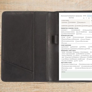 May include: A black leatherette portfolio with a notepad and pen, open to reveal a printed form labeled "Psychotherapist Session Notes." A cup of coffee and a notepad are on the wooden surface.