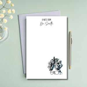 May include: A white notepad with the text "A Note From Dr. Smith" in black script. Below the text is a detailed illustration of an eye exam machine and a pair of eyeglasses. A silver pen rests on the right side.