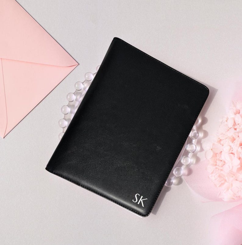 May include: A black leather journal with the initials "SK" in the bottom right corner. The journal is surrounded by a string of clear beads, a pink envelope, and a pink floral arrangement.