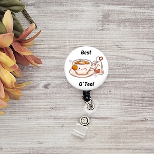 May include: White round badge featuring a cartoon teacup and teabag. The text on the badge reads "Best O' Tea!" with two pink hearts either side. The badge is attached to a black retractable badge reel.