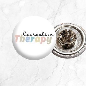 May include: A white button pin with the text "Recreation Therapy" in colorful letters. The pin has a silver back with a safety pin.