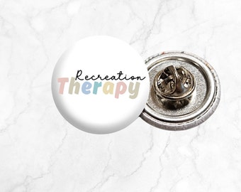 Op de afbeelding: Een witte knoopsluiting met de tekst "Recreation Therapy" in kleurrijke letters. De speld heeft een zilveren achterkant met een veiligheidsspeld.
