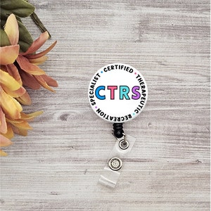 May include: White round badge with the text "Certified Therapeutic Recreation Specialist" and the acronym "CTRS" in colorful letters. The badge is attached to a black retractable badge reel.