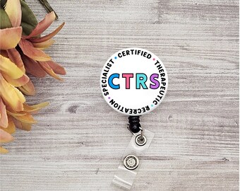 Pode incluir: Crachá redondo branco com o texto "Especialista em Recreação Terapêutica Certificado" e a sigla "CTRS" em letras coloridas. O crachá está preso a um carretel retrátil de crachá preto.