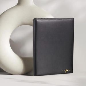 May include: A black leather portfolio with gold initials "jw" in a script font. The portfolio is rectangular and appears to be closed. A cream-colored, abstract ceramic vase is in the background.