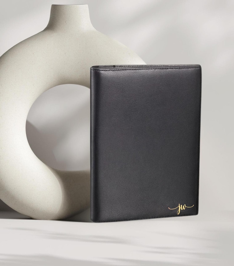 May include: A black leather portfolio with gold initials "jw" in a script font. The portfolio is rectangular and appears to be closed. A cream-colored, abstract ceramic vase is in the background.
