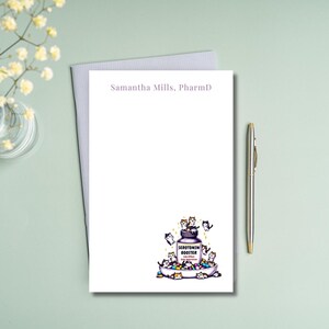 May include: A white notepad with the text "Samantha Mills, PharmD" at the top. The notepad features a cartoon illustration of a bottle of "Serotonin Booster" with a number of cute cats on top of it.