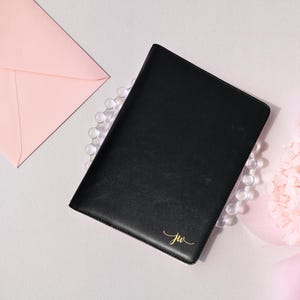 May include: Black leather journal with gold foil initials "jw" in the bottom right corner. A pink envelope and a strand of clear beads are next to the journal. A pink floral arrangement is in the background.