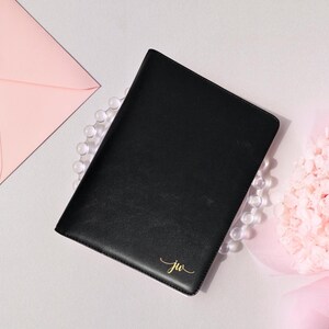 May include: A black leather journal with gold initials "jw" in the corner. The journal is surrounded by a pink envelope, a string of clear beads, and a pink flower arrangement. The journal is closed.