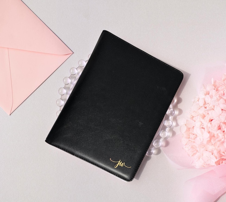 May include: A black leather journal with gold initials "jw" in the corner. The journal is surrounded by a pink envelope, a string of clear beads, and a pink flower arrangement. The journal is closed.