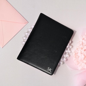 May include: A black leather journal with the initials "SK" in the bottom right corner. The journal is surrounded by a string of clear beads, a pink envelope, and a bouquet of pink flowers.