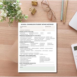May include: A white notepad titled "SCHOOL COUNSELOR STUDENT INTAKE NOTEPAD" lies on a wooden desk, surrounded by office supplies. The notepad features checkboxes for student information, concerns, strengths, and academic history. A calculator, laptop, and glasses are also visible.
