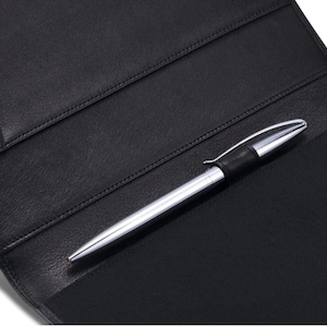 May include: A black leather notebook or portfolio with interior pockets. A silver pen with a black grip rests inside the open notebook. The pen has a silver tip and a black band near the top.