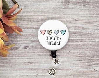 Pode incluir: Carretel de crachá branco com um botão branco que apresenta uma fileira de quatro corações coloridos e o texto "RECREATION THERAPIST".
