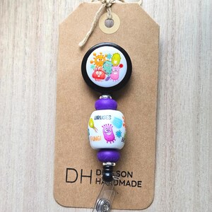 May include: A retractable badge reel with a cartoon virus design. The reel features a black top with colorful virus illustrations, a purple center section with the words "VIRUSES" and "FUNGI", and a clear plastic clip. The reel is attached to a brown tag.