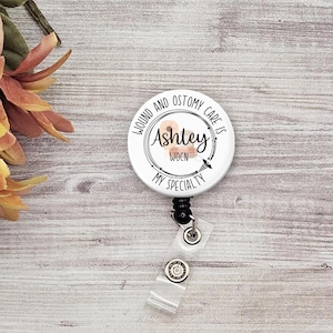 May include: White round badge holder with a black retractable cord. The badge features a white background with black text that reads "Wound and Ostomy Care is my Specialty". The text is surrounded by a black circle with a white outline. The name "Ashley" is written in black text inside the circle. The letters "WOCN" are written in black text below the name. A small graphic of a bandage is inside the circle.