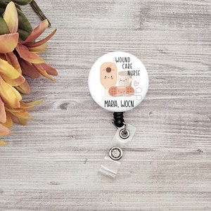 May include: A white badge reel with a cartoon illustration of medical supplies, including a bandage, scissors, and a bottle. The text "Wound Care Nurse" and "Maria, WOCN" are printed on the badge. The reel has a clear plastic strap and a black retractable cord.